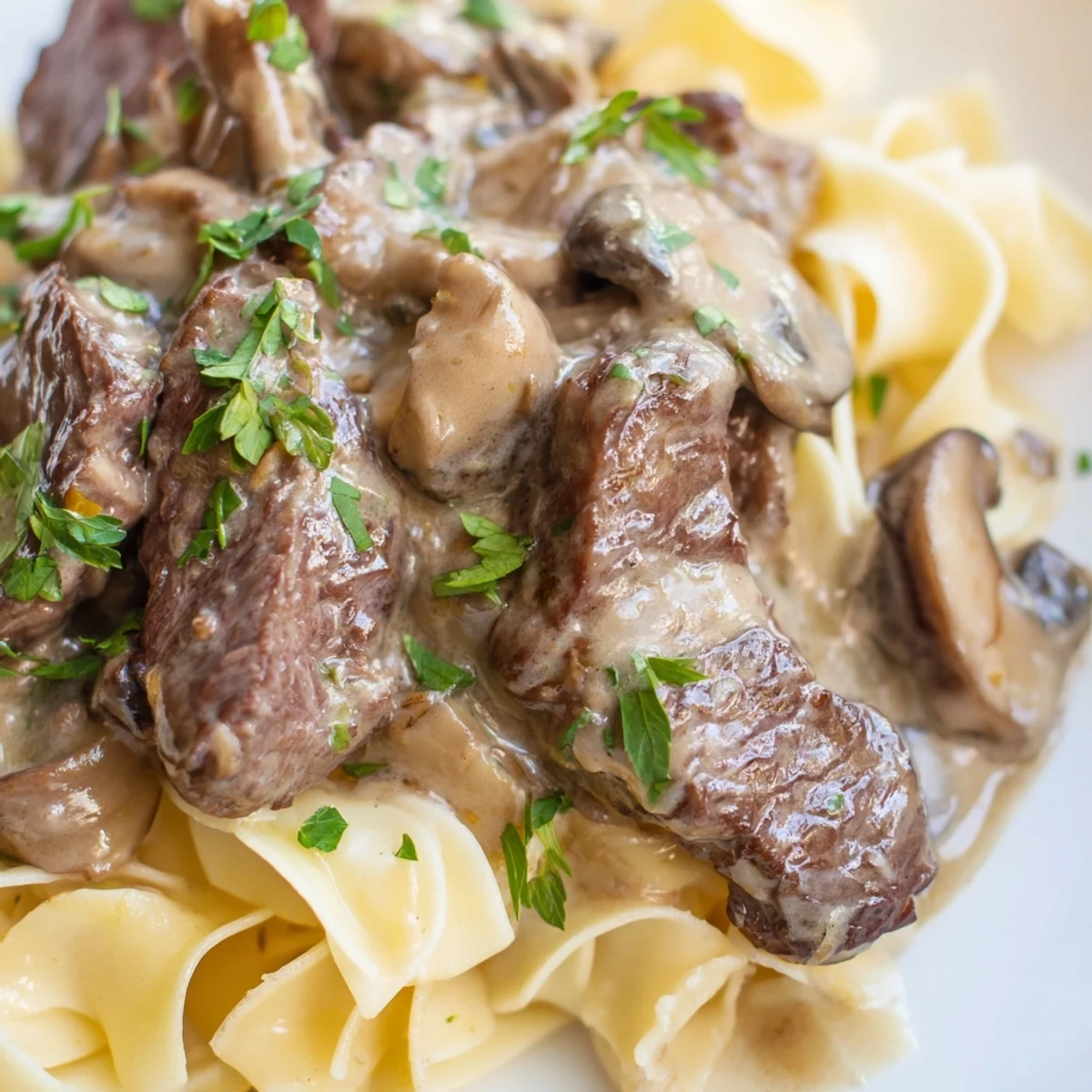 Golden, creamy Beef and Mushroom Stroganoff coats tender egg noodles, garnished with fresh parsley in a white bowl.