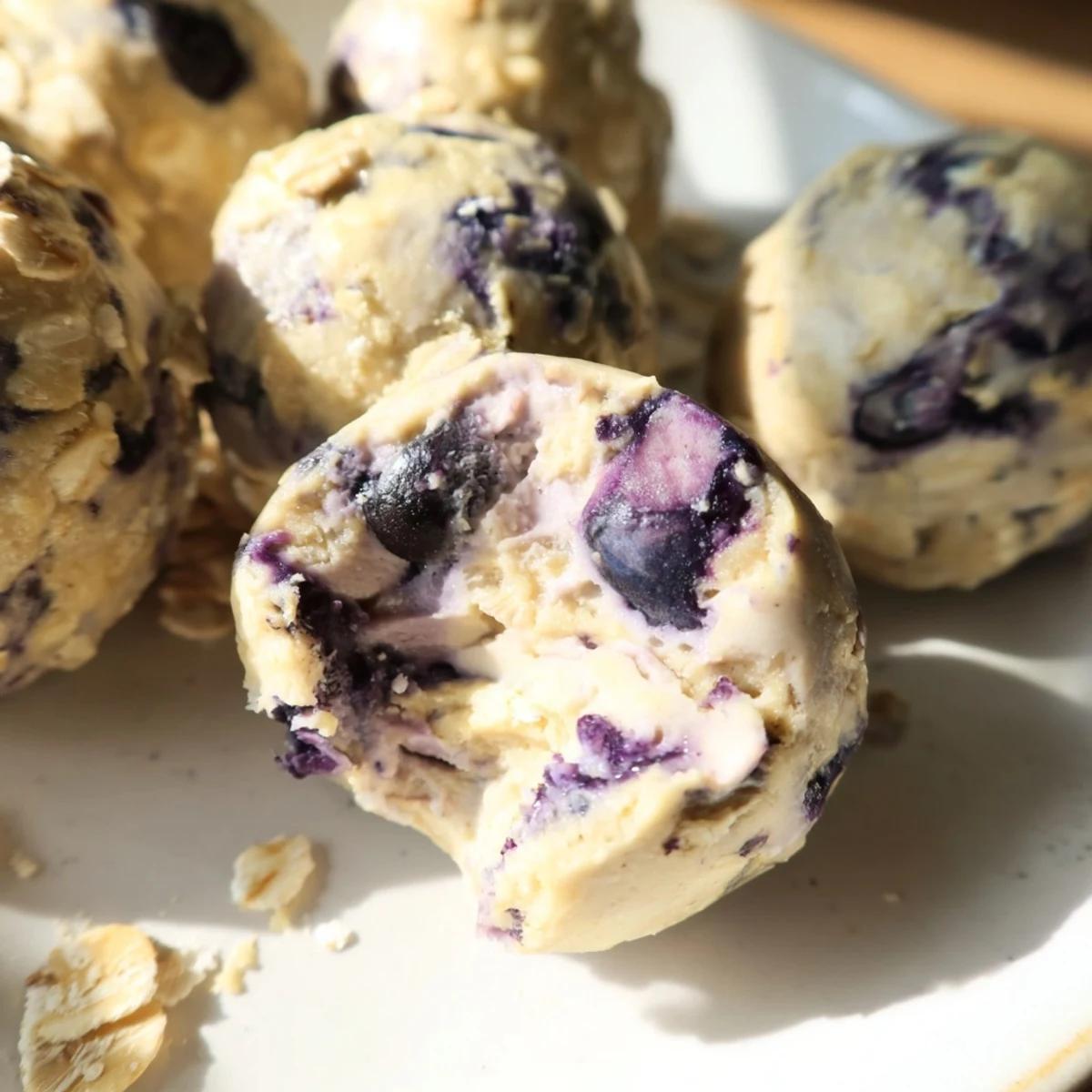 A close-up of Blueberry Cheesecake Protein Bites on a wooden board, showing lemon zest flecks and blueberry bits, served alongside a glass of milk.