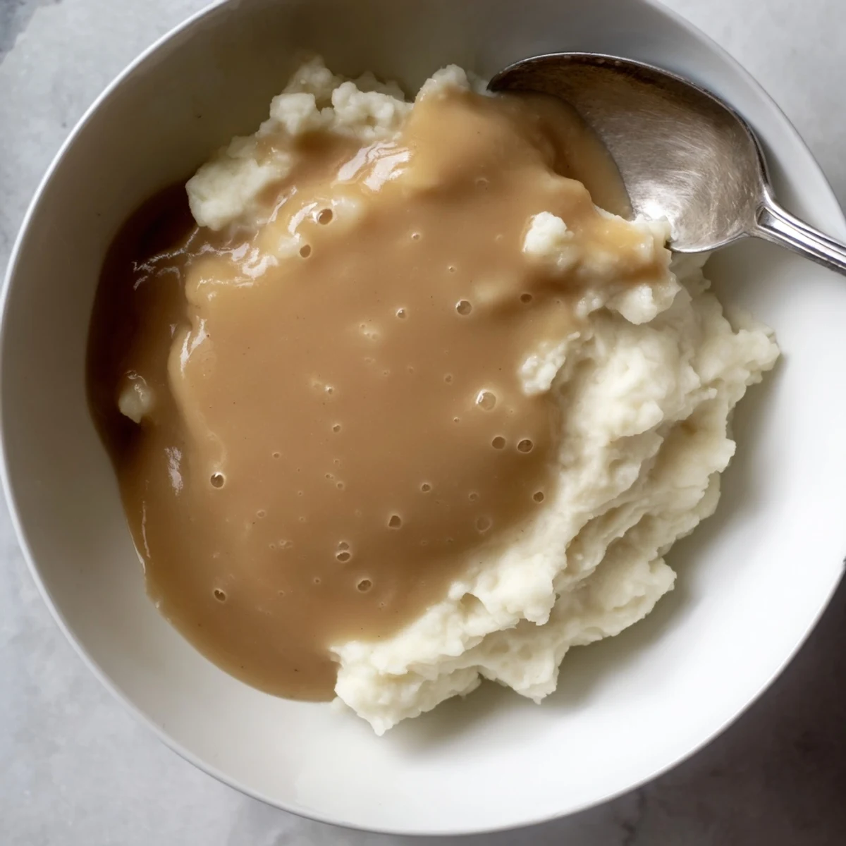 Golden homemade gravy poured from a whisk over rustic mashed potatoes in a white bowl
