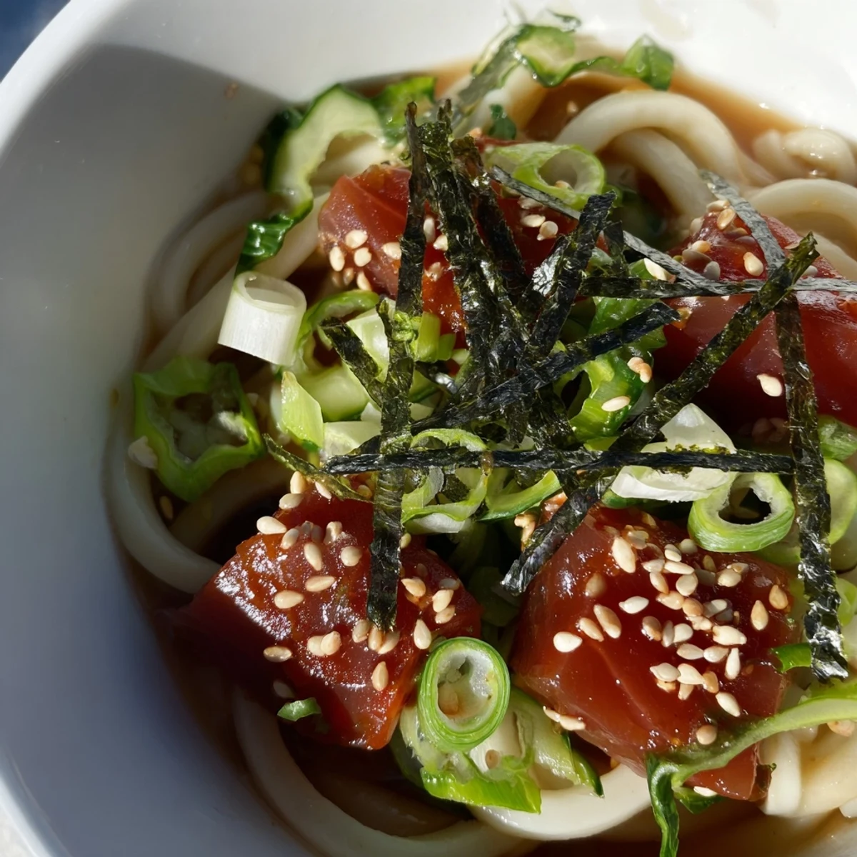 Bowl of spicy tuna udon noodles topped with sesame seeds, green onions, and fresh cucumber