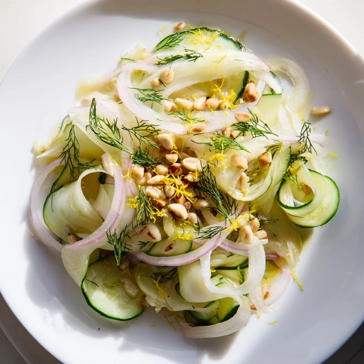 Golden pine nuts garnish this crisp fennel cucumber salad on a white plate.